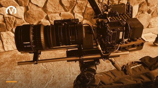 A cinema camera with a Canon 50 to 1000mm zoom lens on a tripod, set up on location with camera cases on the ground behind it.